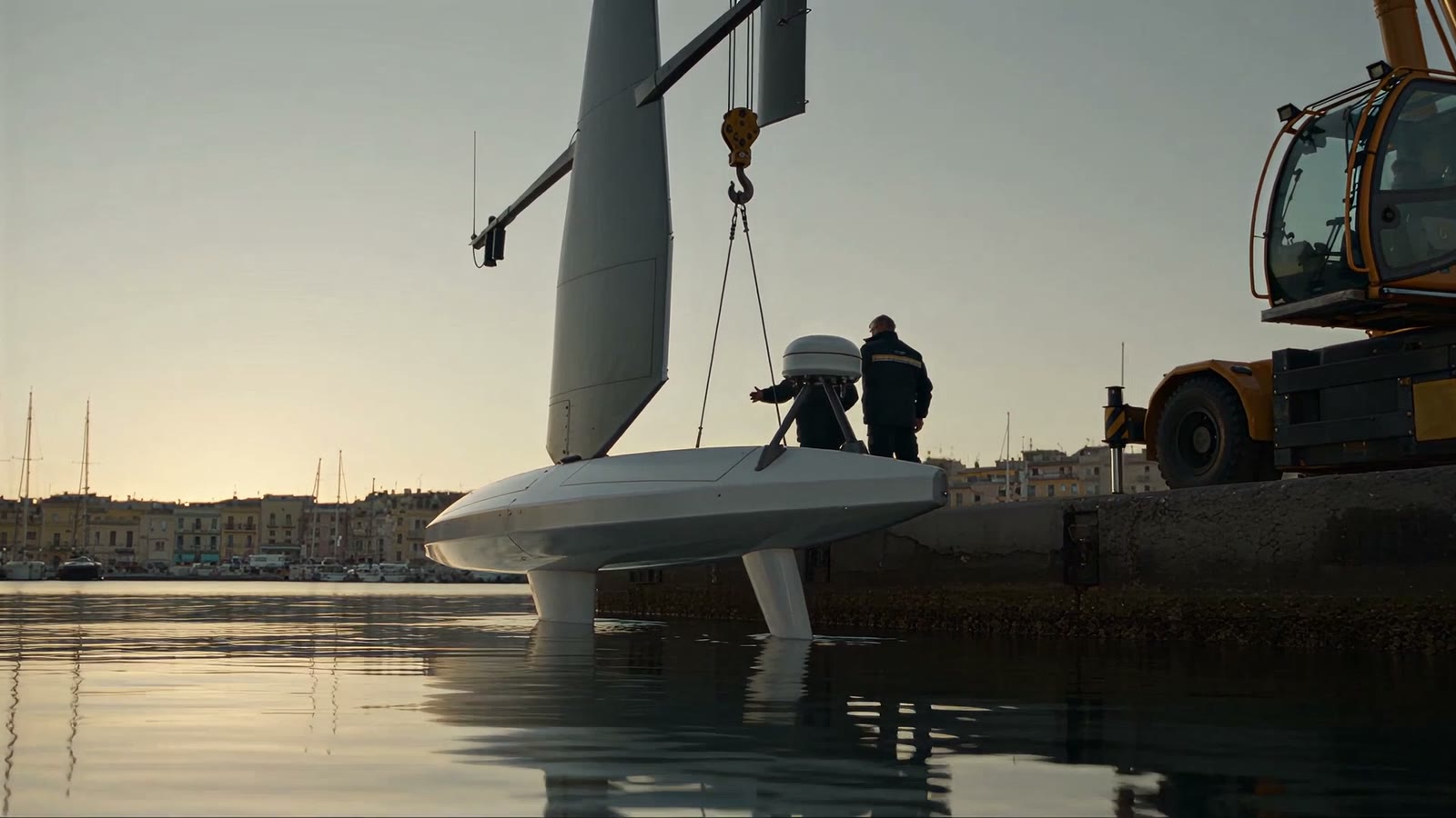 A crane lowers the sail drone into the harbour at dawn while two technicians steady it